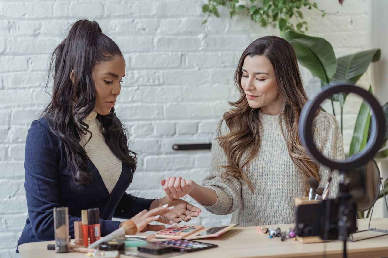 Jeunes créatrices de contenu positives et d’origines diverses testant des produits de maquillage, assises à une table et enregistrant un vlog sur un smartphone posé sur un trépied avec anneau lumineux
